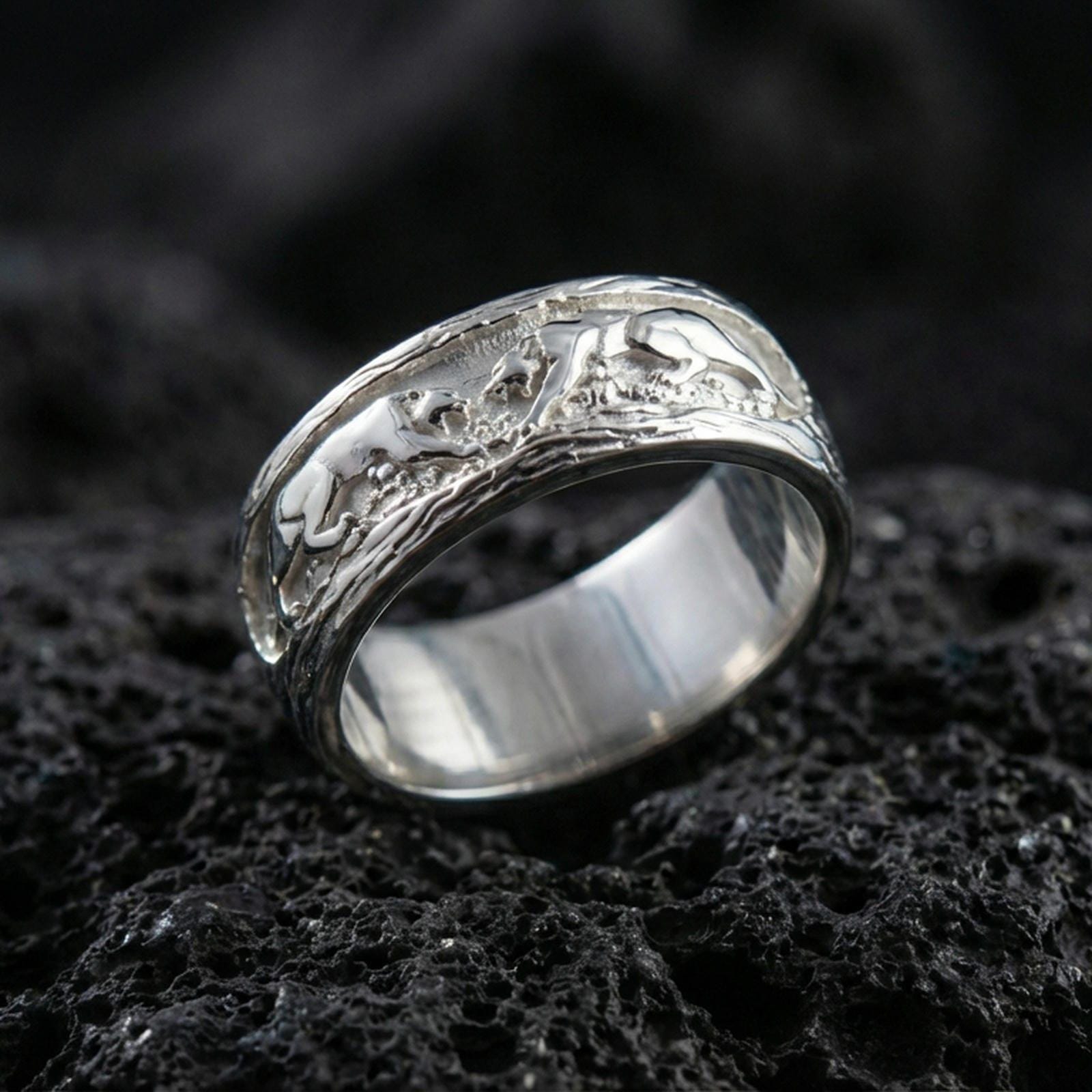 Sterling silver band ring photographed on black volcanic stone, shown in a polished silver finish with crisp highlights across the curved edges and sculpted figures.