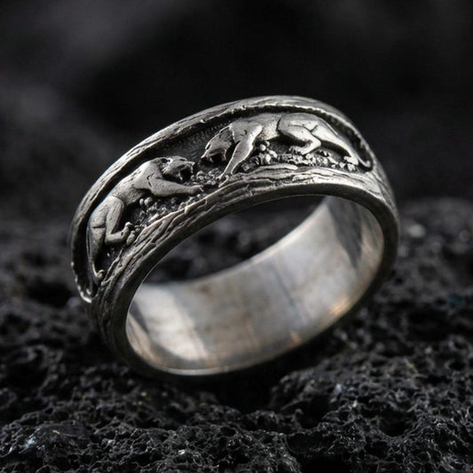Sterling silver band ring photographed on black volcanic stone, featuring an oxidized finish that brings out the raised three-monkey relief and shadowed details.