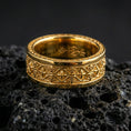 Load image into Gallery viewer, Gothic fleur-de-lis band ring on black volcanic stone, shown in a gold plated finish with warm shine highlighting the ornate cross-and-lily motifs.
