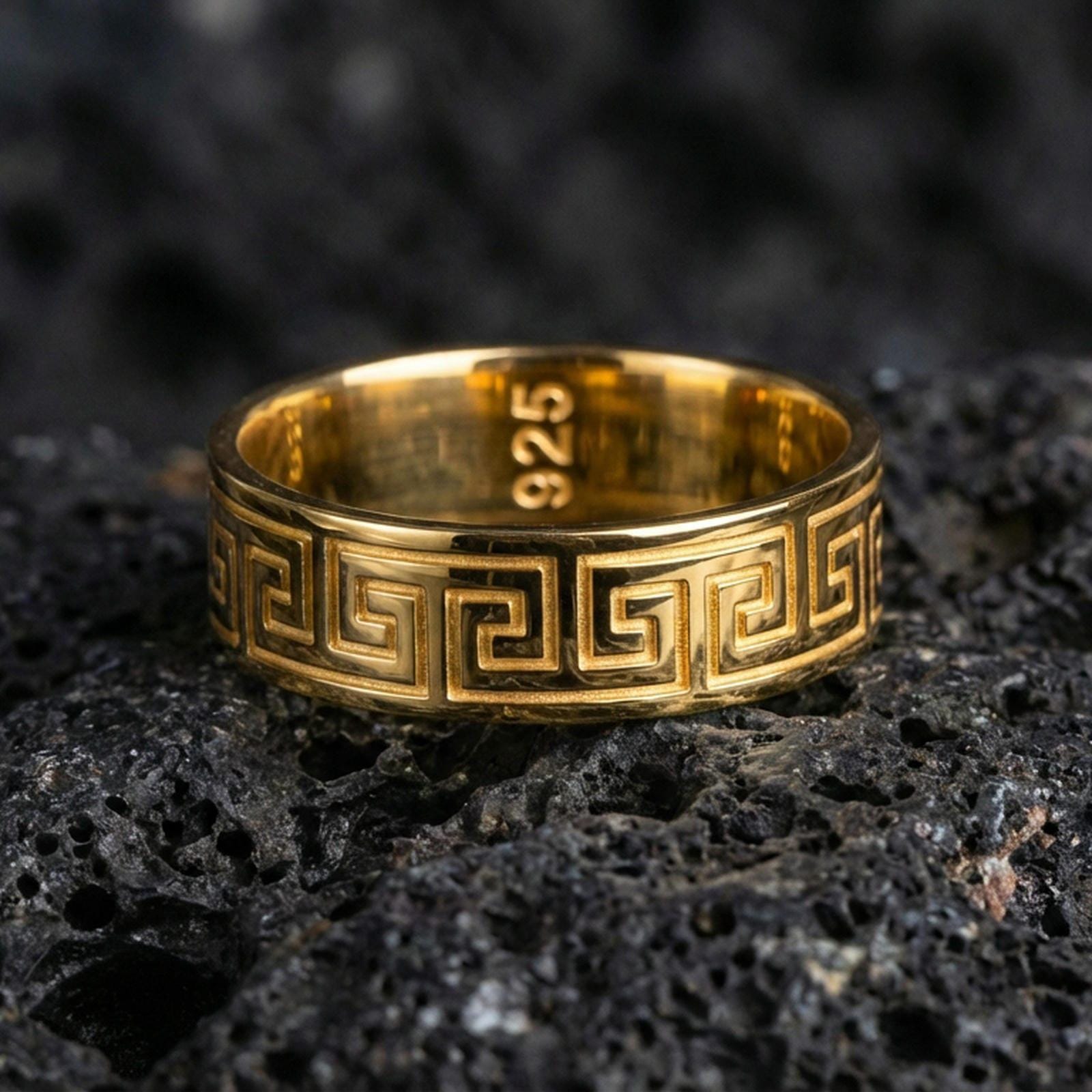 Gold plated Greek key pattern band ring on volcanic rock, classic meander motif with crisp edge detail, luxury jewelry product photography.