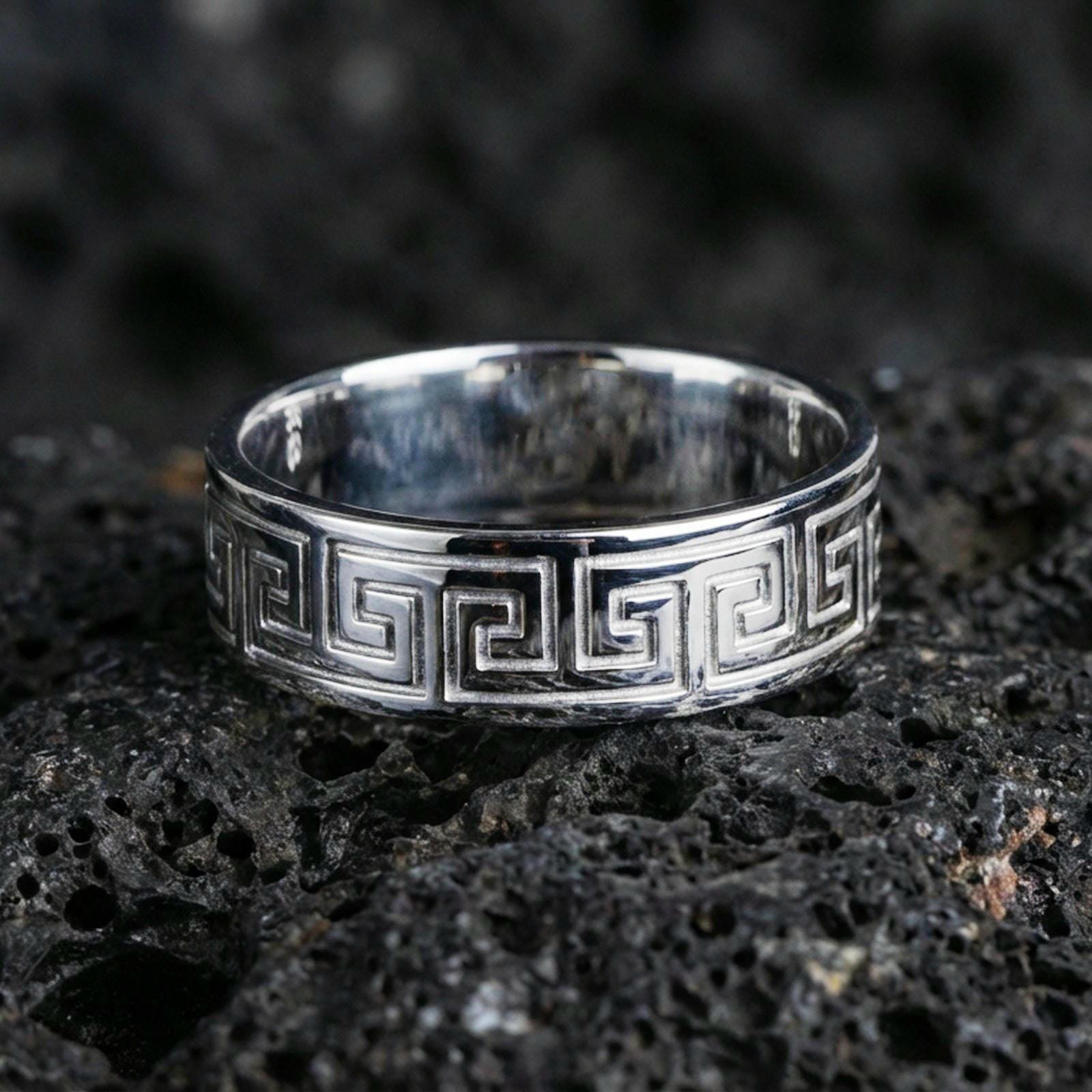Polished silver Greek key wedding band on black volcanic stone, clean geometric meander border, centered close-up studio lighting.