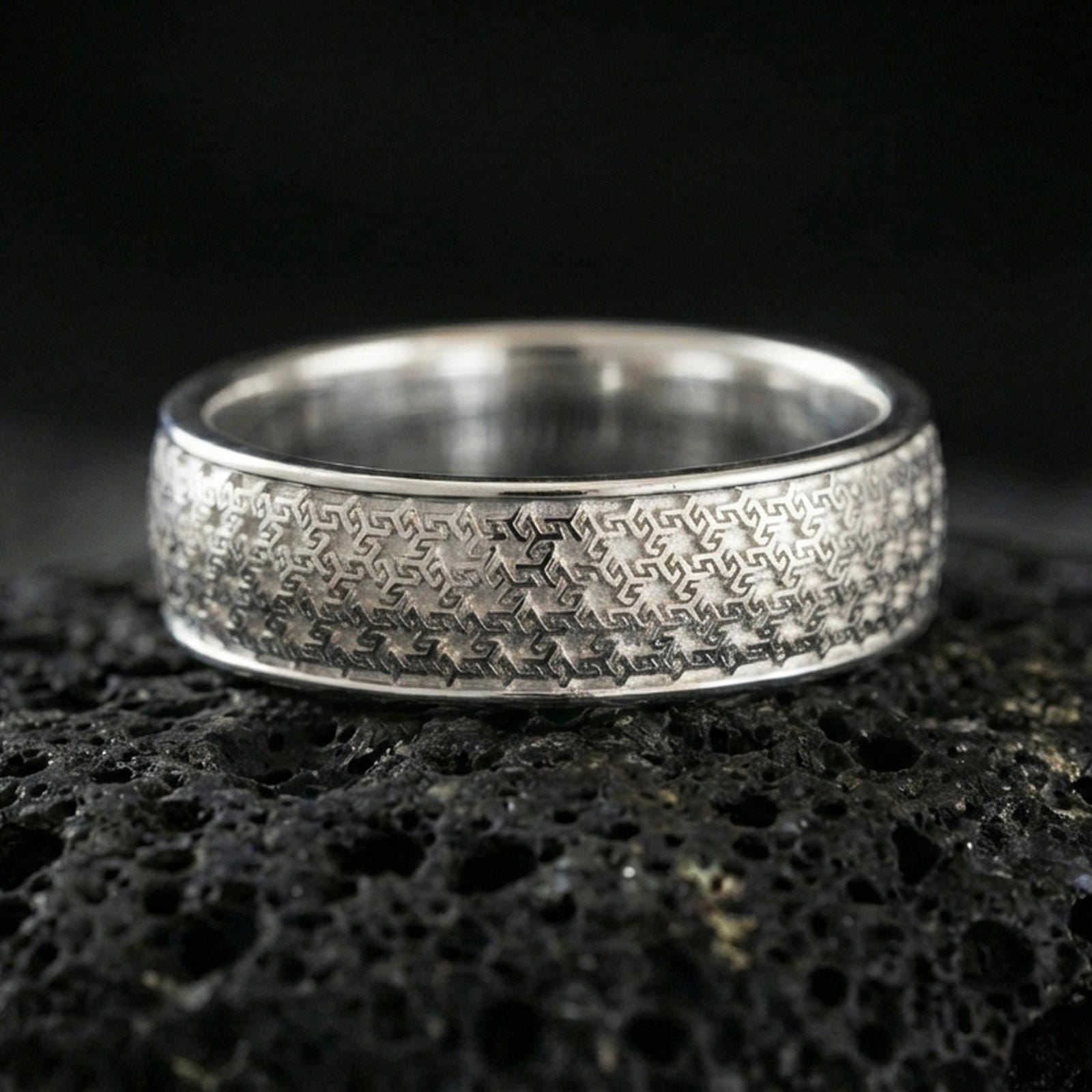 Polished silver textured band ring on black volcanic stone, crisp interwoven geometric pattern engraving, centered close-up studio lighting.