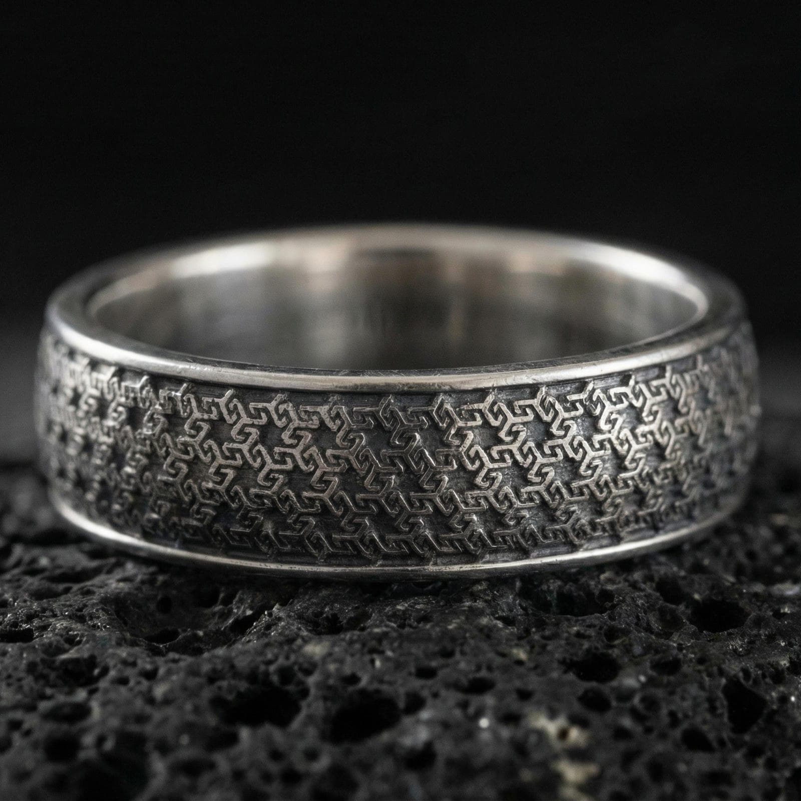 Oxidized sterling silver geometric band ring on dark volcanic rock, repeating interlocking maze texture, premium macro product shot.