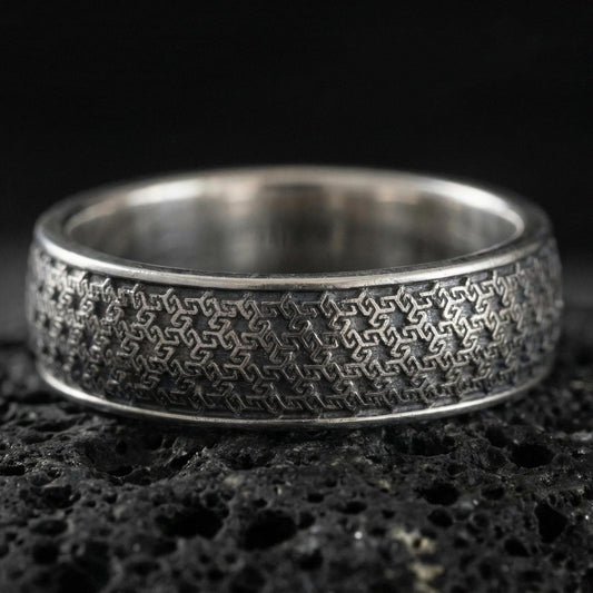 Oxidized sterling silver geometric band ring on dark volcanic rock, repeating interlocking maze texture, premium macro product shot.