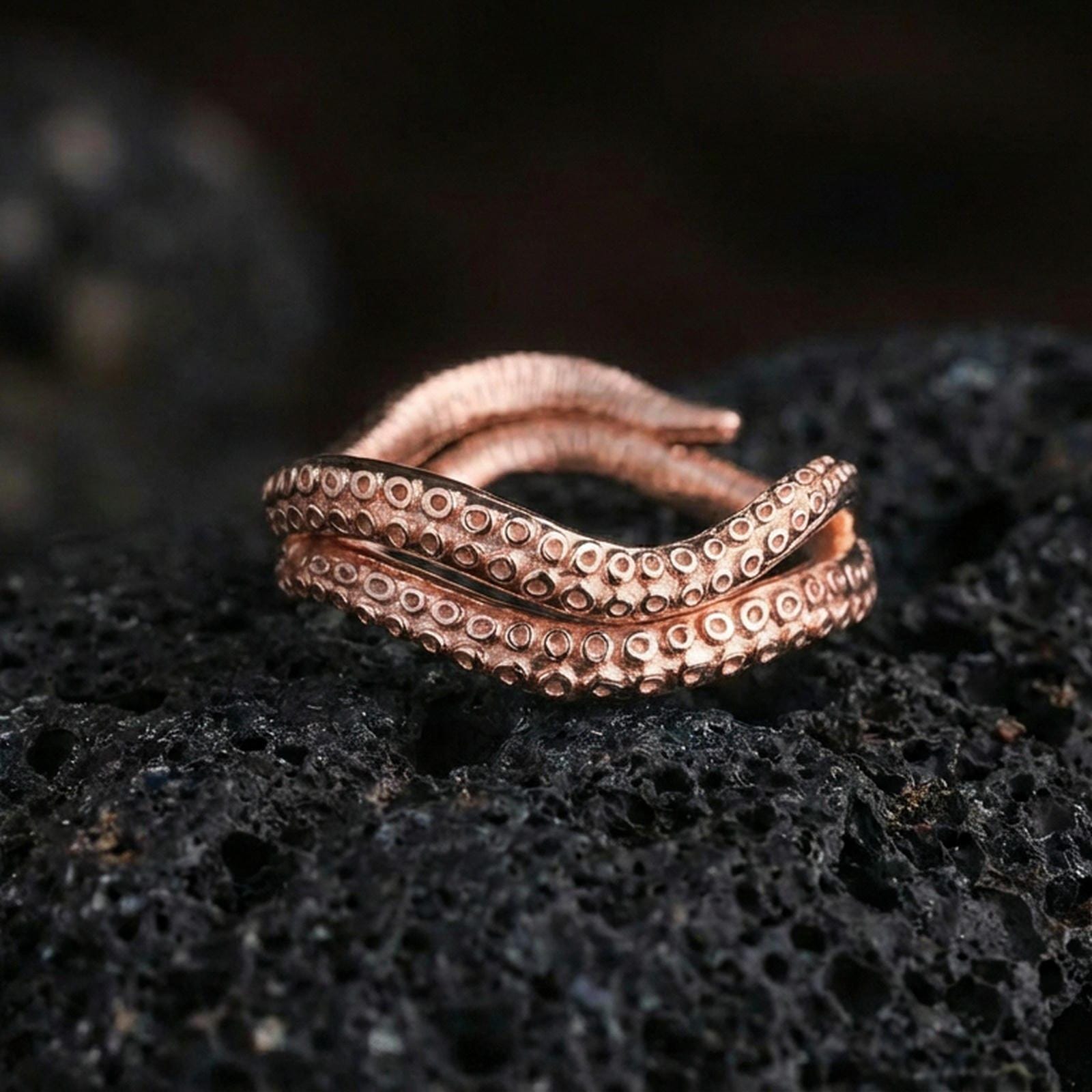 Rose gold plated sterling silver octopus tentacle ring on volcanic stone, sculpted suction cup texture with deep contrast, minimal high-end close-up.