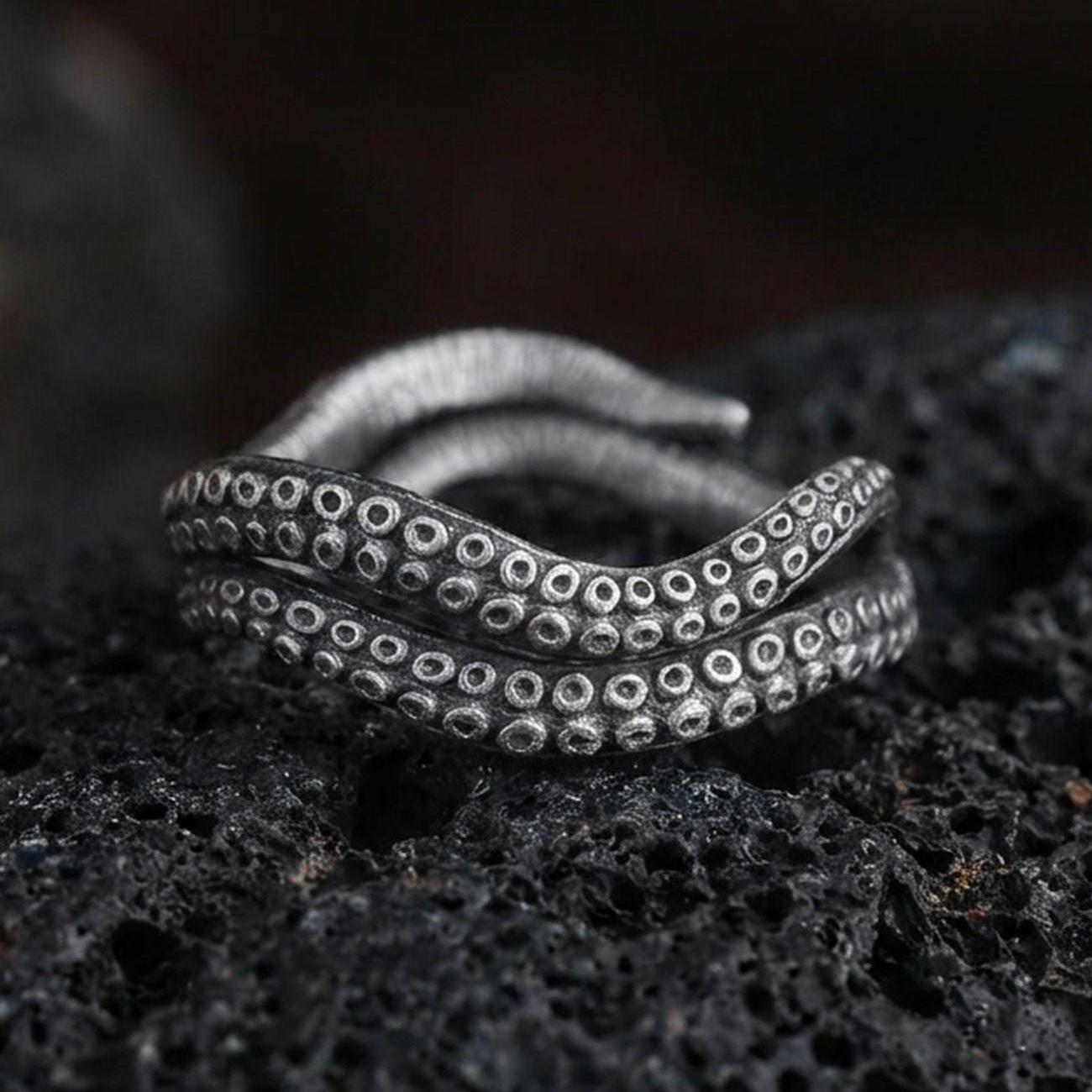 Oxidized sterling silver octopus tentacle wrap ring on dark volcanic rock, suction cup texture detail, premium macro product shot.