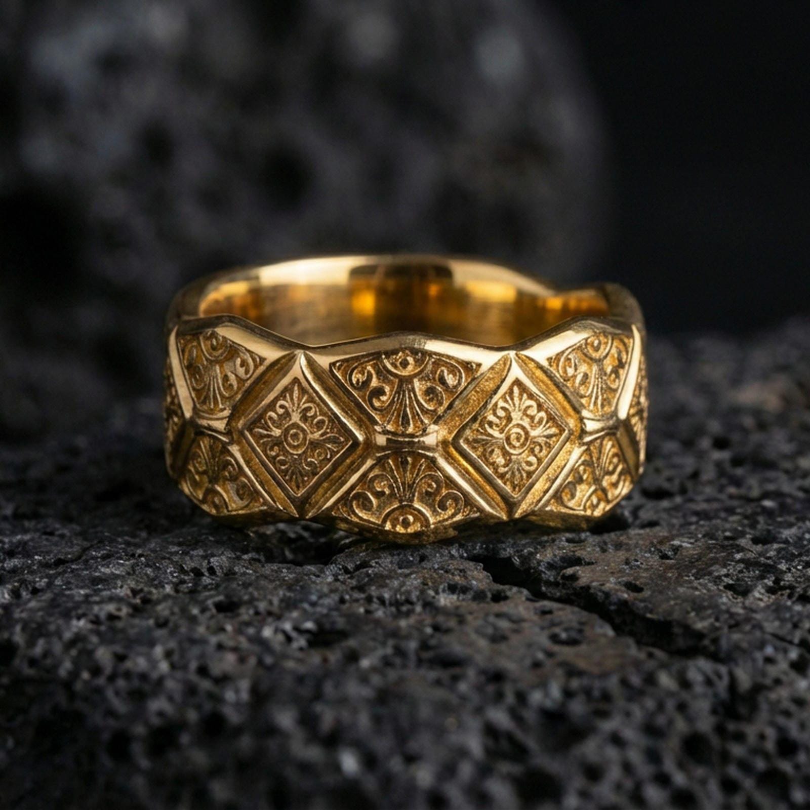 Gold plated ornate engraved band ring on volcanic rock, raised diamond pattern with decorative scroll accents, luxury jewelry product photography.