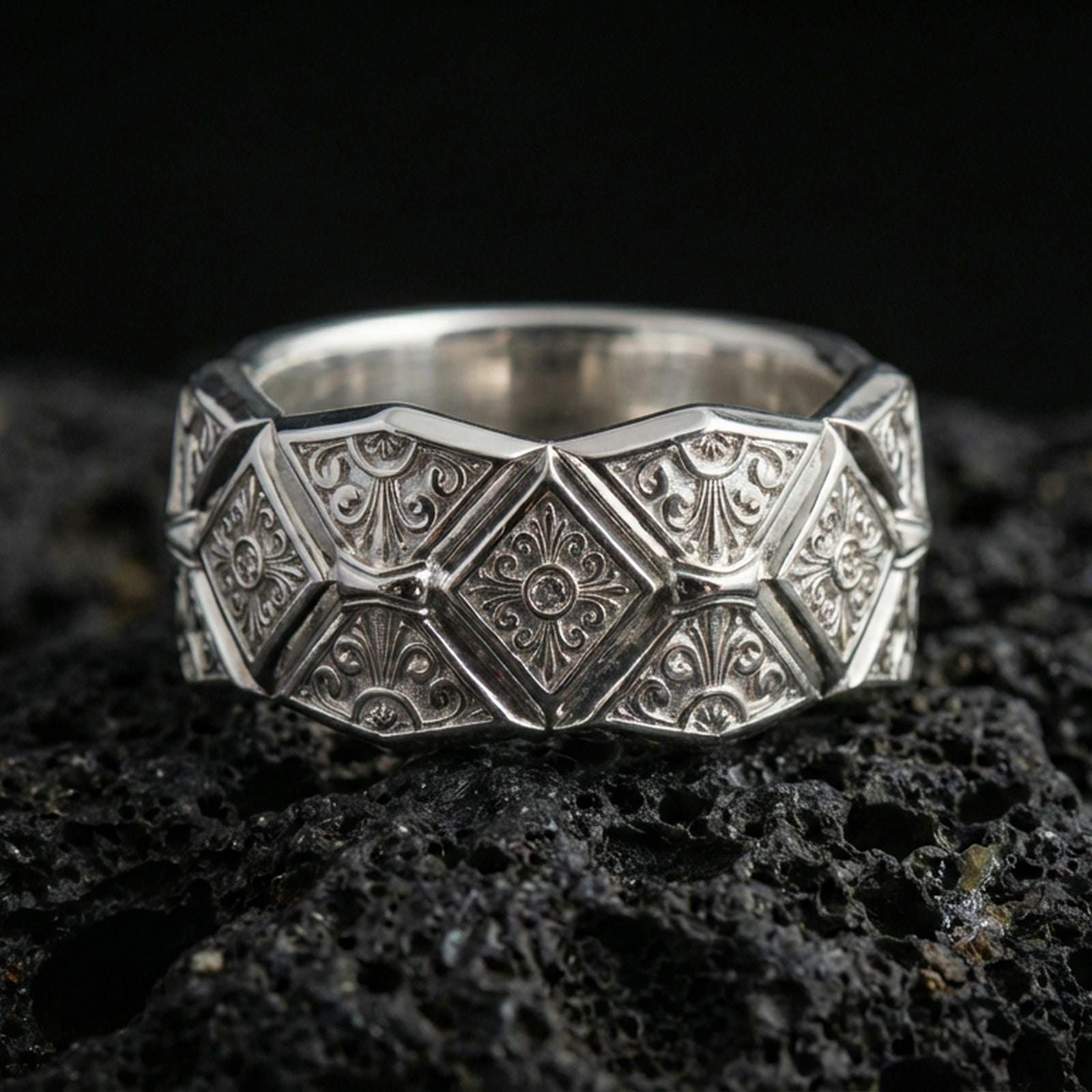 Polished silver geometric engraved band ring on black volcanic stone, crisp faceted diamond motifs and ornate scroll engraving, centered close-up studio lighting.