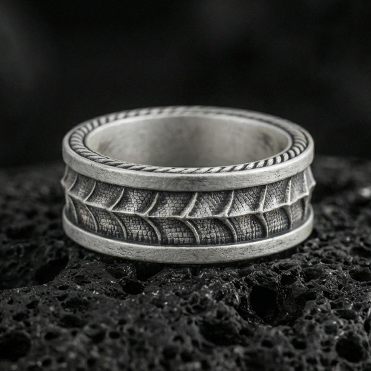 Oxidized sterling silver serpent skin band ring on dark volcanic rock, scaled texture detail with rope edge border, premium macro product shot.