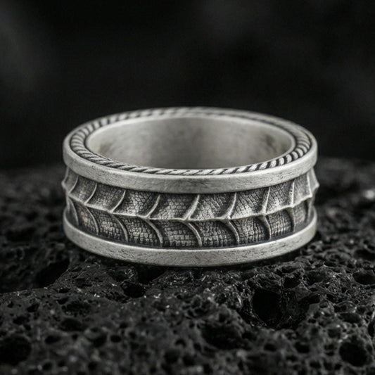 Oxidized sterling silver serpent skin band ring on dark volcanic rock, scaled texture detail with rope edge border, premium macro product shot.