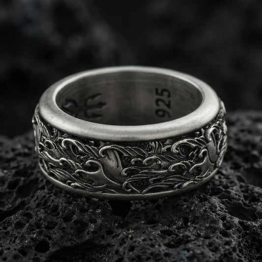 Oxidized sterling silver spinning wave band ring on dark volcanic rock, nautical ocean swirl engraving with bold relief detail, premium macro product shot.