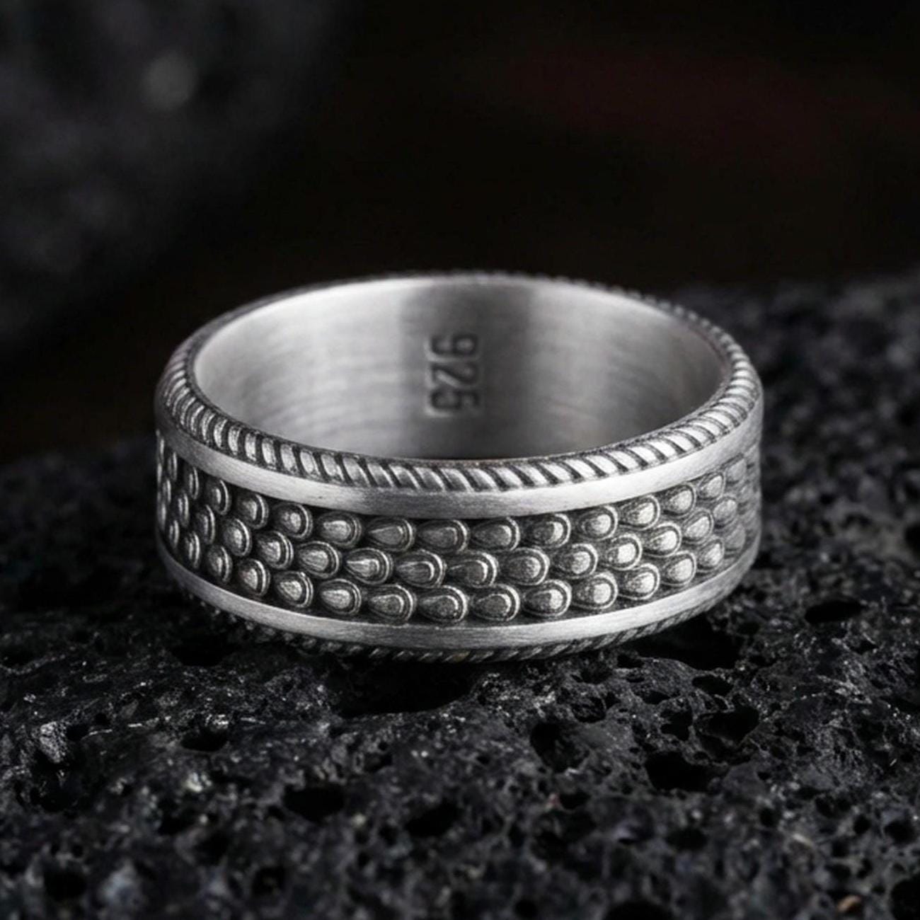 Oxidized sterling silver beaded dot texture band ring on dark volcanic rock, hammered bubble pattern with rope edge border, premium macro product shot.
