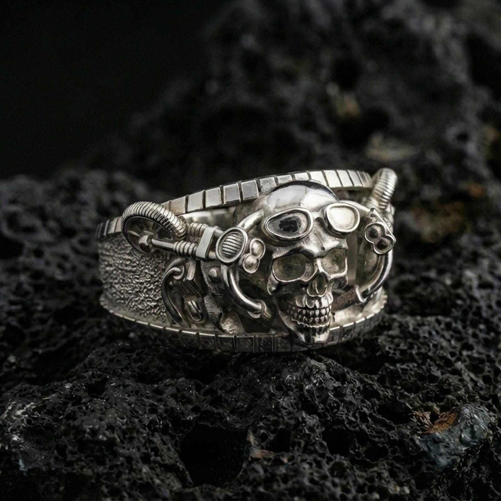 Polished silver steampunk skull statement ring on black volcanic stone, crisp skull and gear-inspired details with textured band, centered close-up studio lighting.