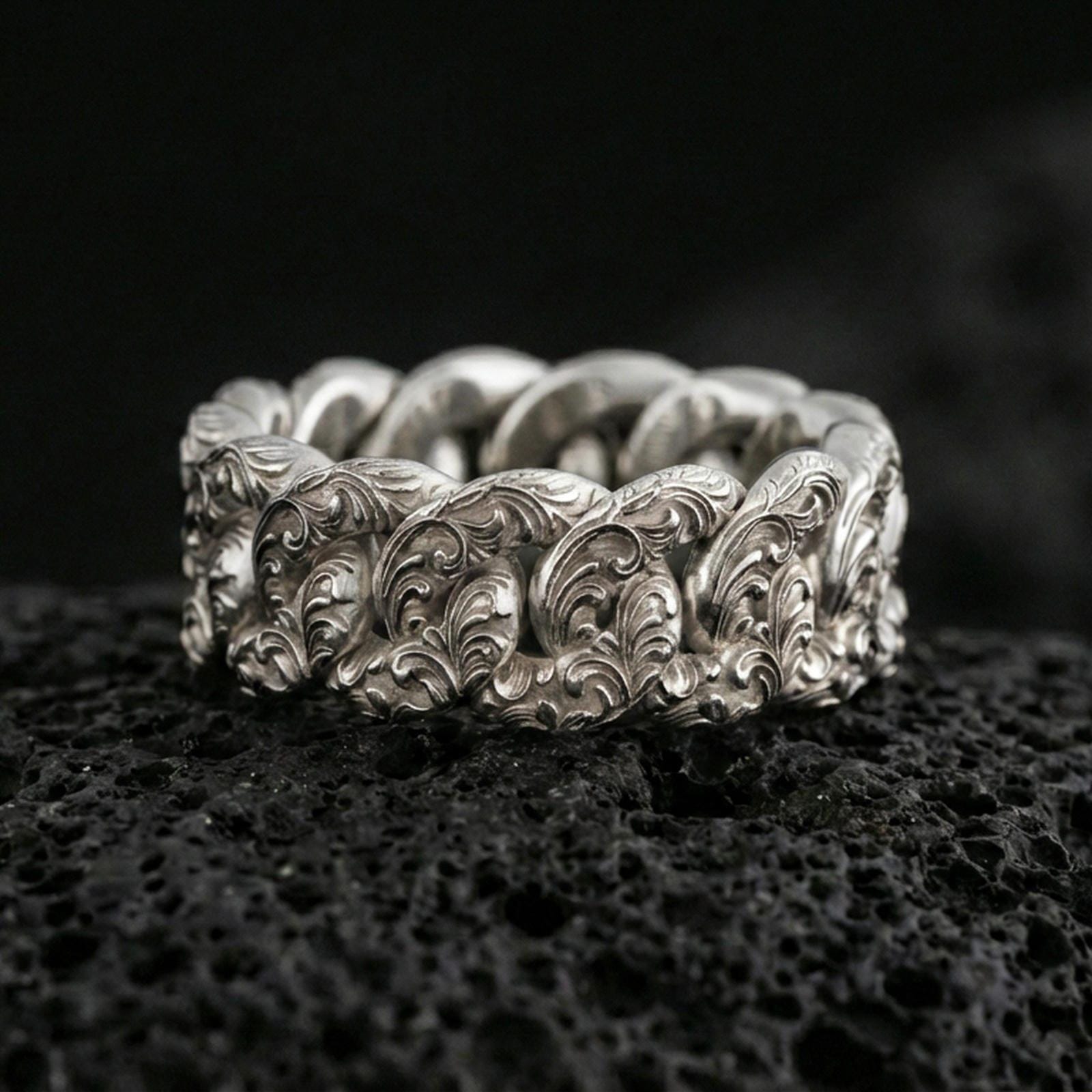 Polished silver Cuban link band ring on black volcanic stone, crisp sculpted chain links with ornate engraved texture, centered close-up studio lighting.