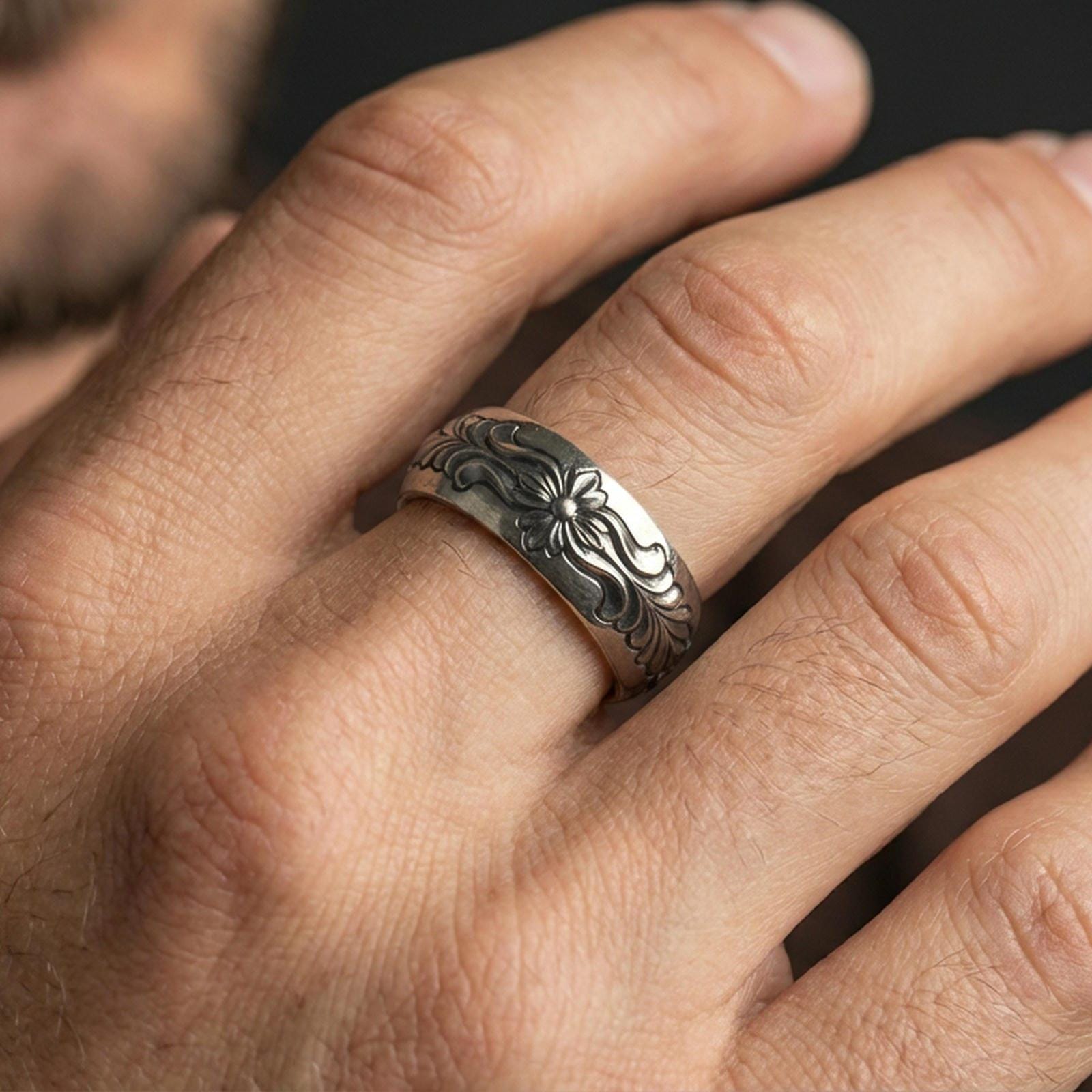 Oxidized sterling silver floral scroll band worn on finger, raised flower detail with flowing leaf engraving, natural lifestyle hand shot with shallow depth of field.
