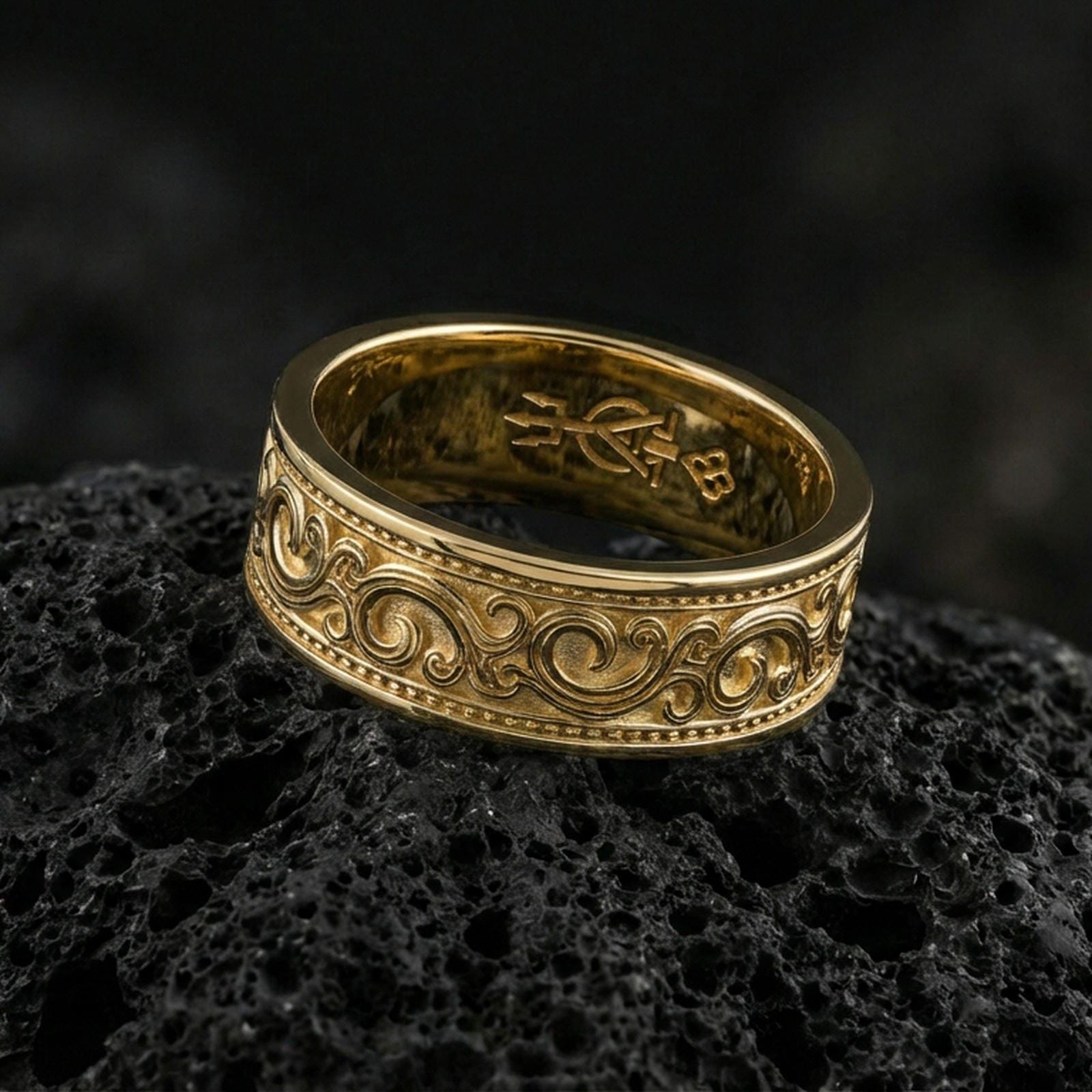 Gold plated scrollwork engraved band ring on volcanic rock, detailed swirl motif with beaded border trim, luxury jewelry product photography.