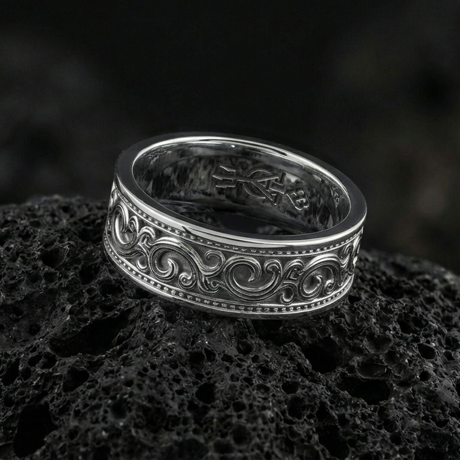 Polished silver engraved scroll band ring on black volcanic stone, crisp swirling filigree detail and dotted edge accents, centered close-up studio lighting.