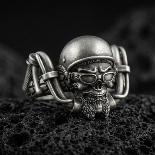 Oxidized sterling silver biker skull ring on dark volcanic rock, helmet and goggles relief with detailed beard, premium macro product shot.