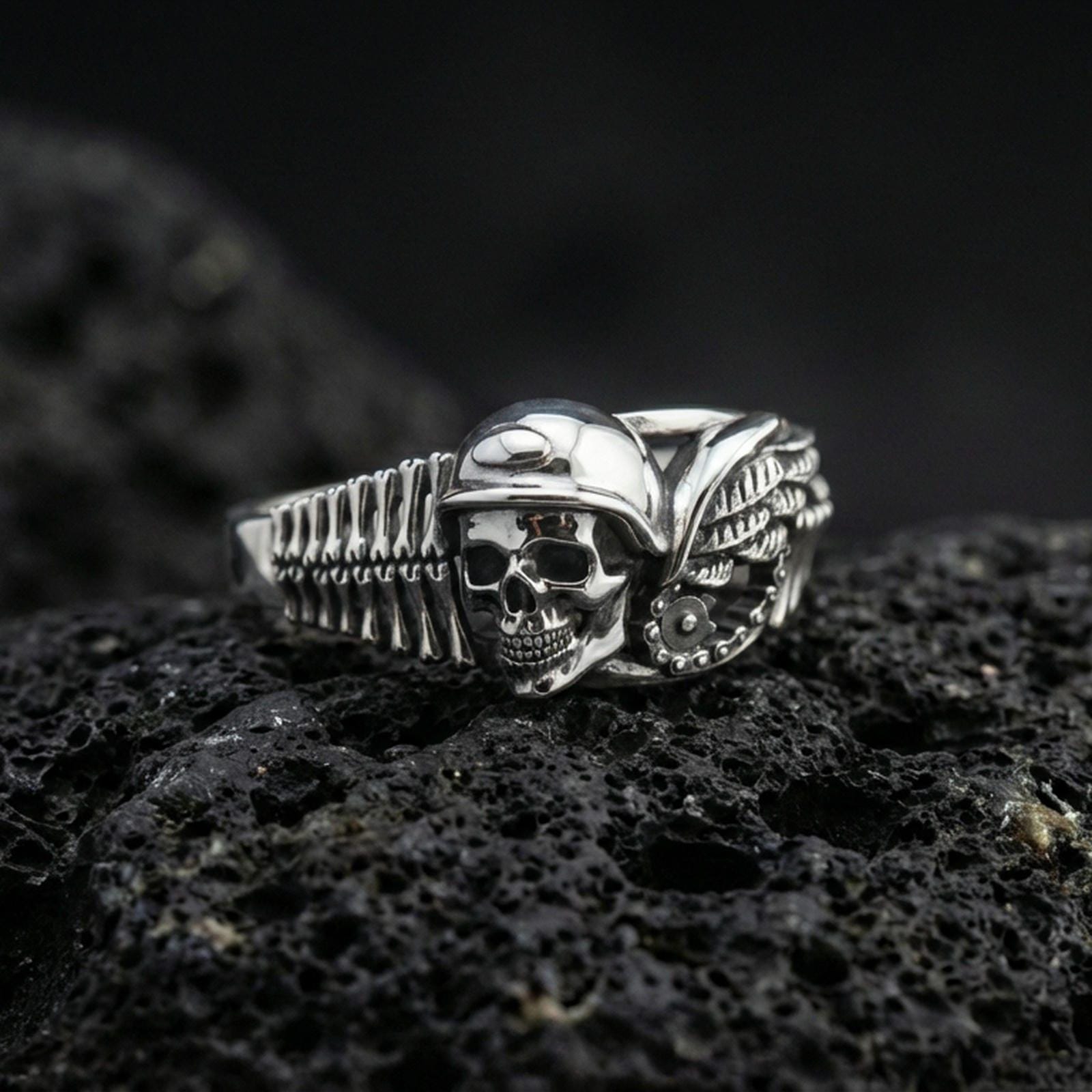 Polished silver gothic biker skull statement ring on black volcanic stone, crisp helmet skull with mechanical wing accents, centered close-up studio lighting.