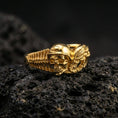 Load image into Gallery viewer, Gold plated biker skull ring on volcanic rock, detailed helmeted skull with engine-inspired wing motif, luxury jewelry product photography.
