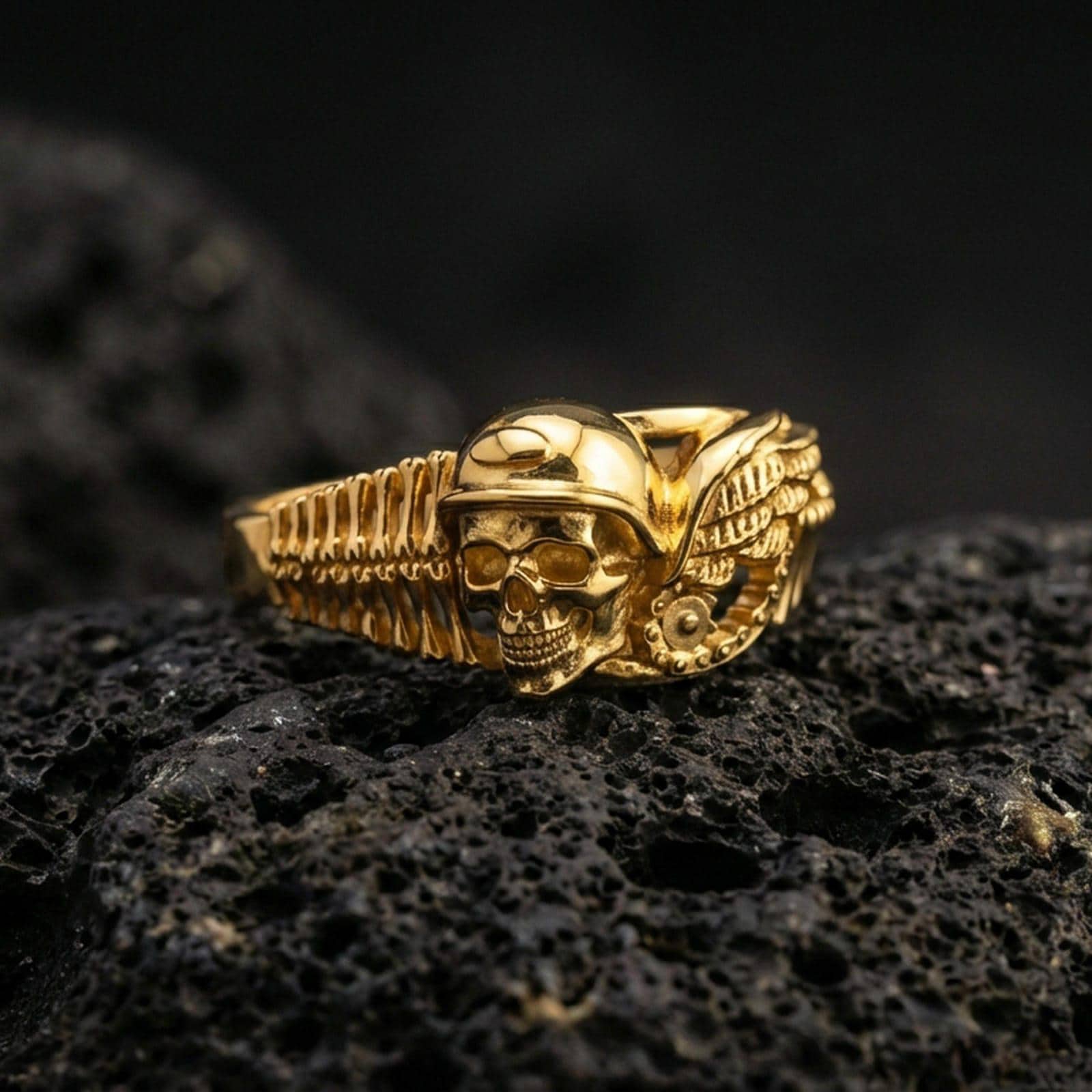 Gold plated biker skull ring on volcanic rock, detailed helmeted skull with engine-inspired wing motif, luxury jewelry product photography.