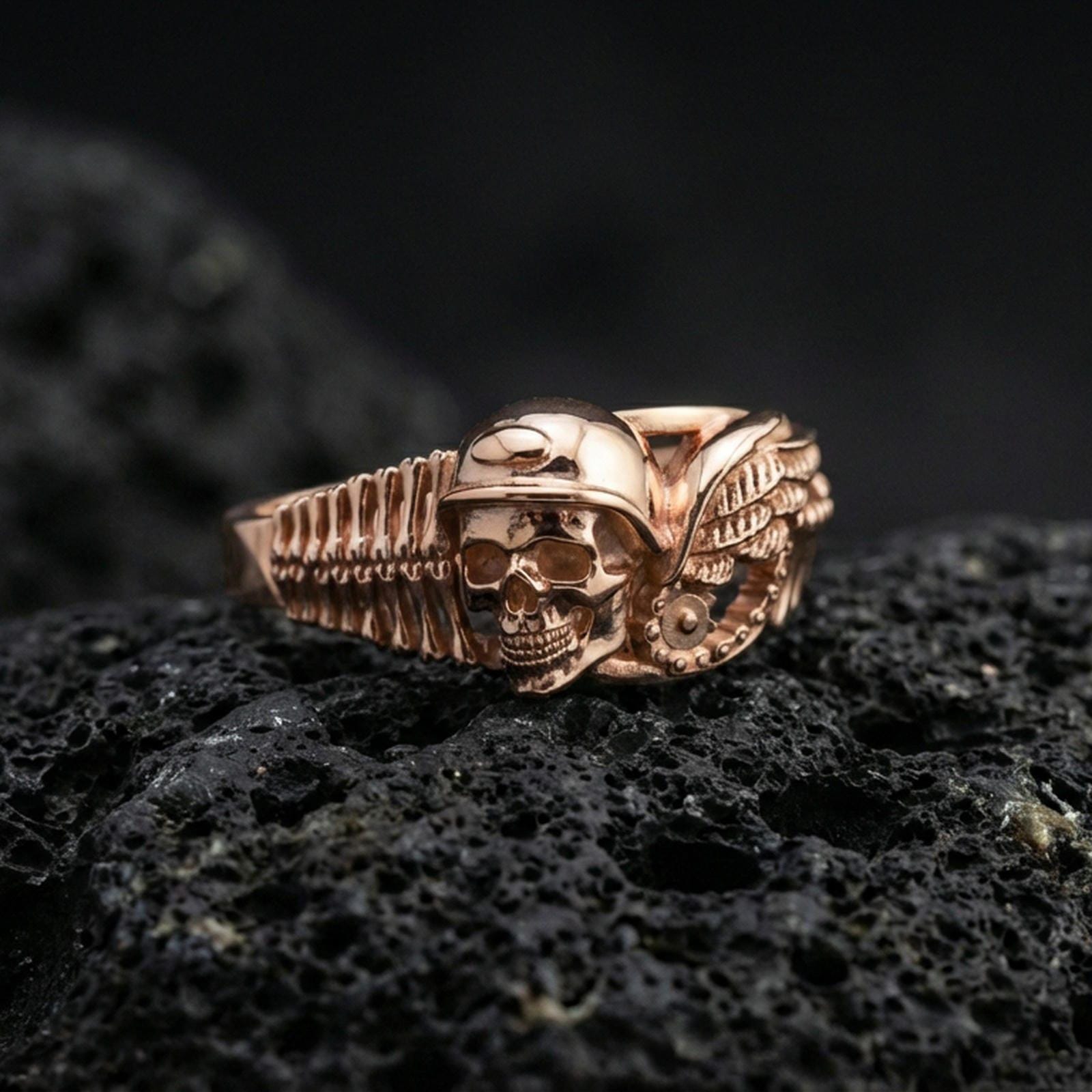 Rose gold plated sterling silver gothic biker skull ring on volcanic stone, sculpted helmeted skull relief with wing detail and deep contrast, minimal high-end close-up.