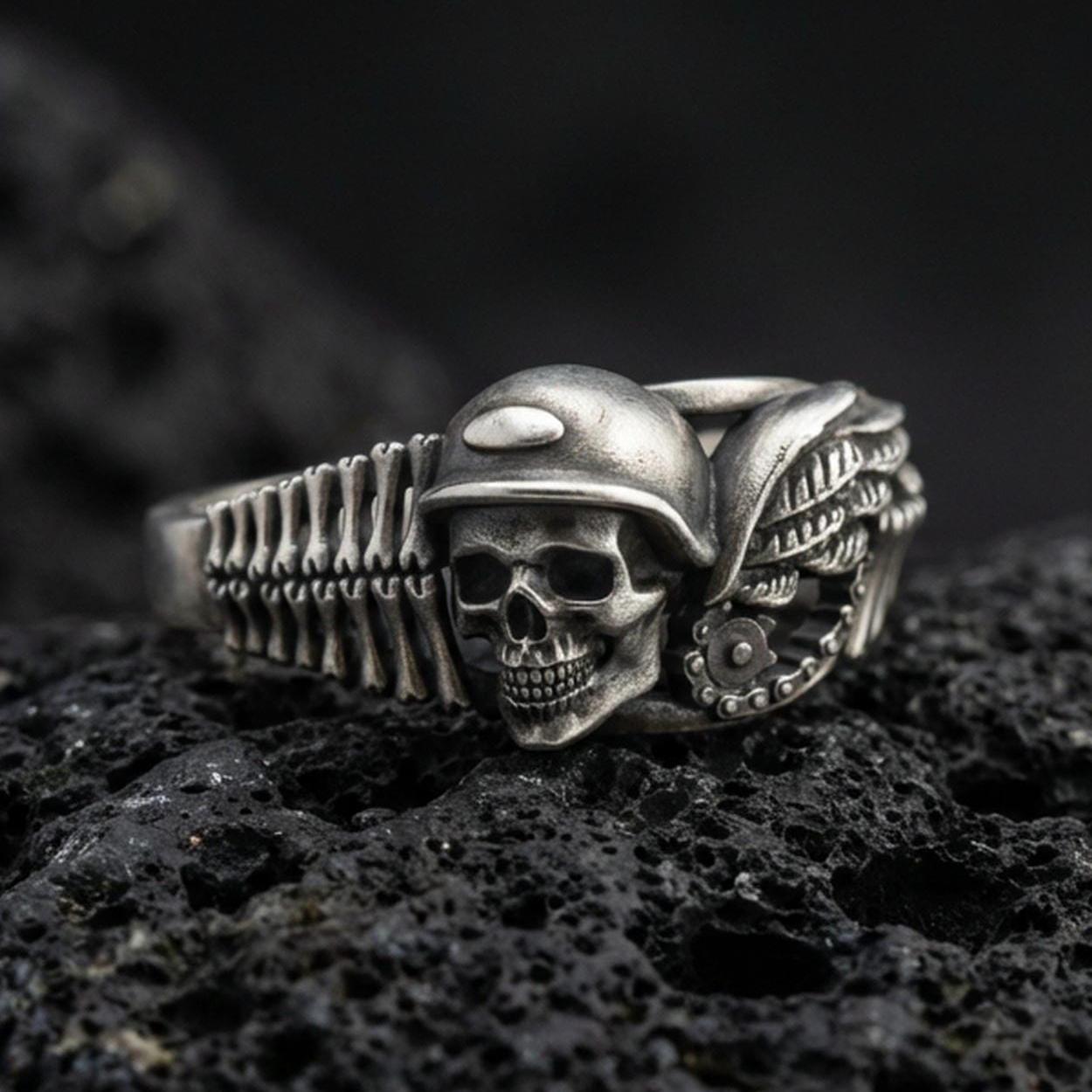 Oxidized sterling silver biker skull ring on dark volcanic rock, helmeted skull relief with wing and engine detail, premium macro product shot.
