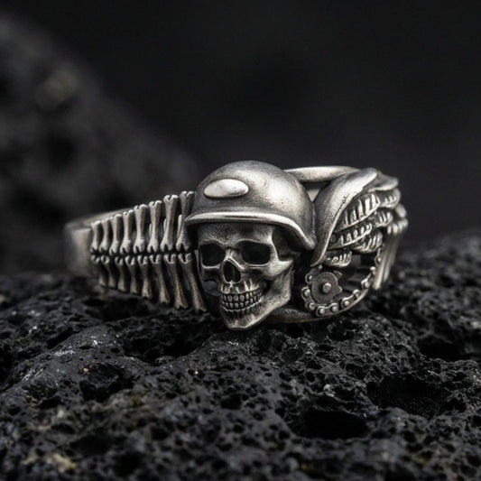 Oxidized sterling silver biker skull ring on dark volcanic rock, helmeted skull relief with wing and engine detail, premium macro product shot.