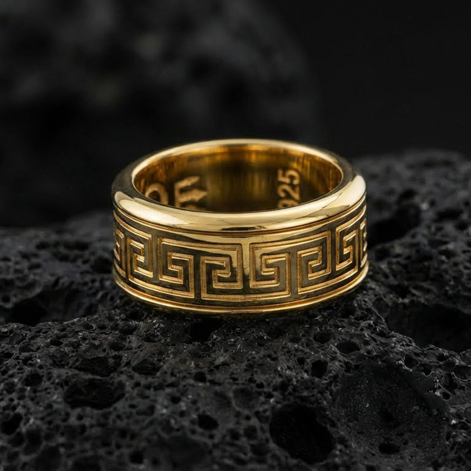 Gold plated Greek key pattern band ring on volcanic rock, classic meander motif with clean engraved lines, luxury jewelry product photography.