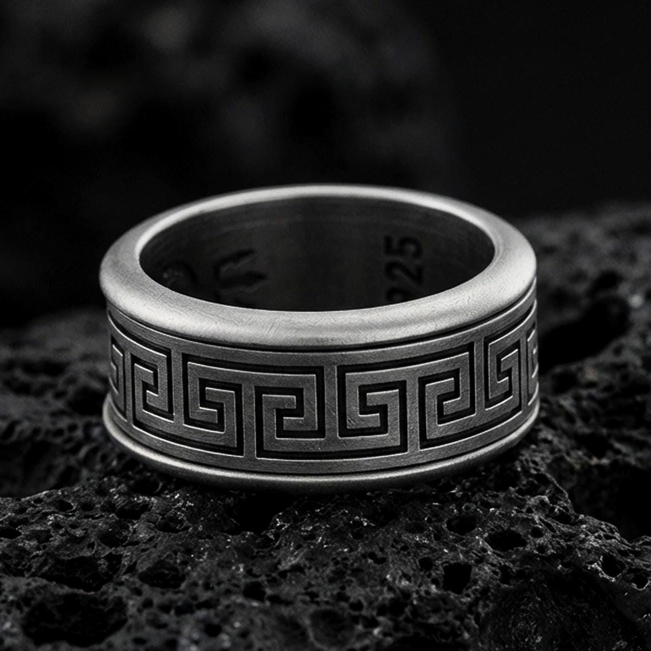 Oxidized sterling silver Greek key band ring on dark volcanic rock, bold meander pattern engraving with smooth comfort edges, premium macro product shot.