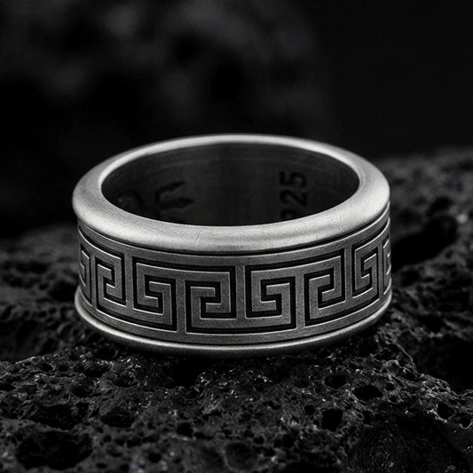 Oxidized sterling silver Greek key band ring on dark volcanic rock, bold meander pattern engraving with smooth comfort edges, premium macro product shot.