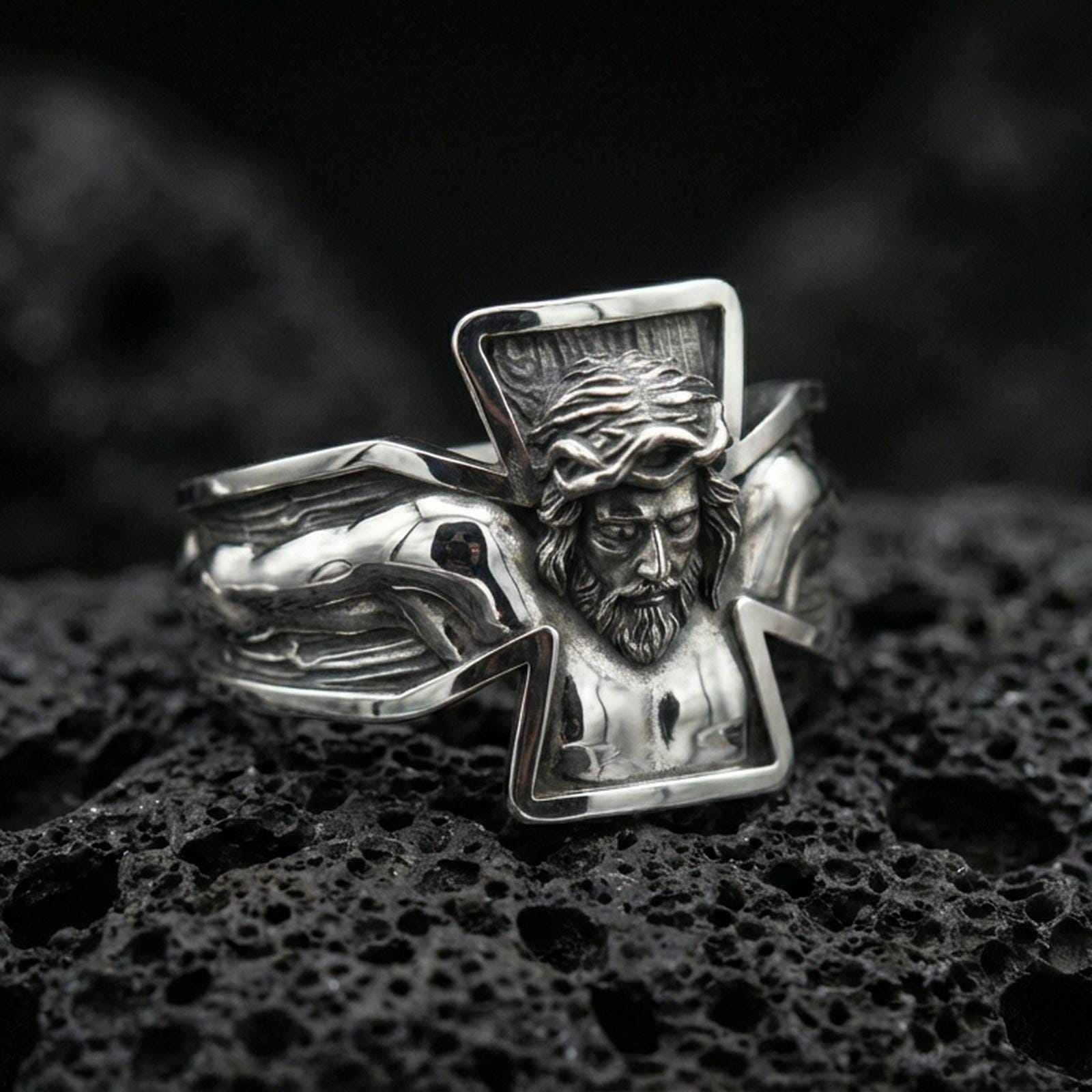 Polished silver Jesus crucifix ring on black volcanic stone, crisp sculpted Christ figure and angular cross outline, centered close-up studio lighting.