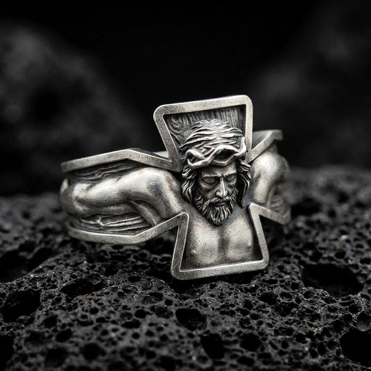 Oxidized sterling silver crucifix Jesus ring on dark volcanic rock, high-relief Christ figure with open cross frame, premium macro product shot.