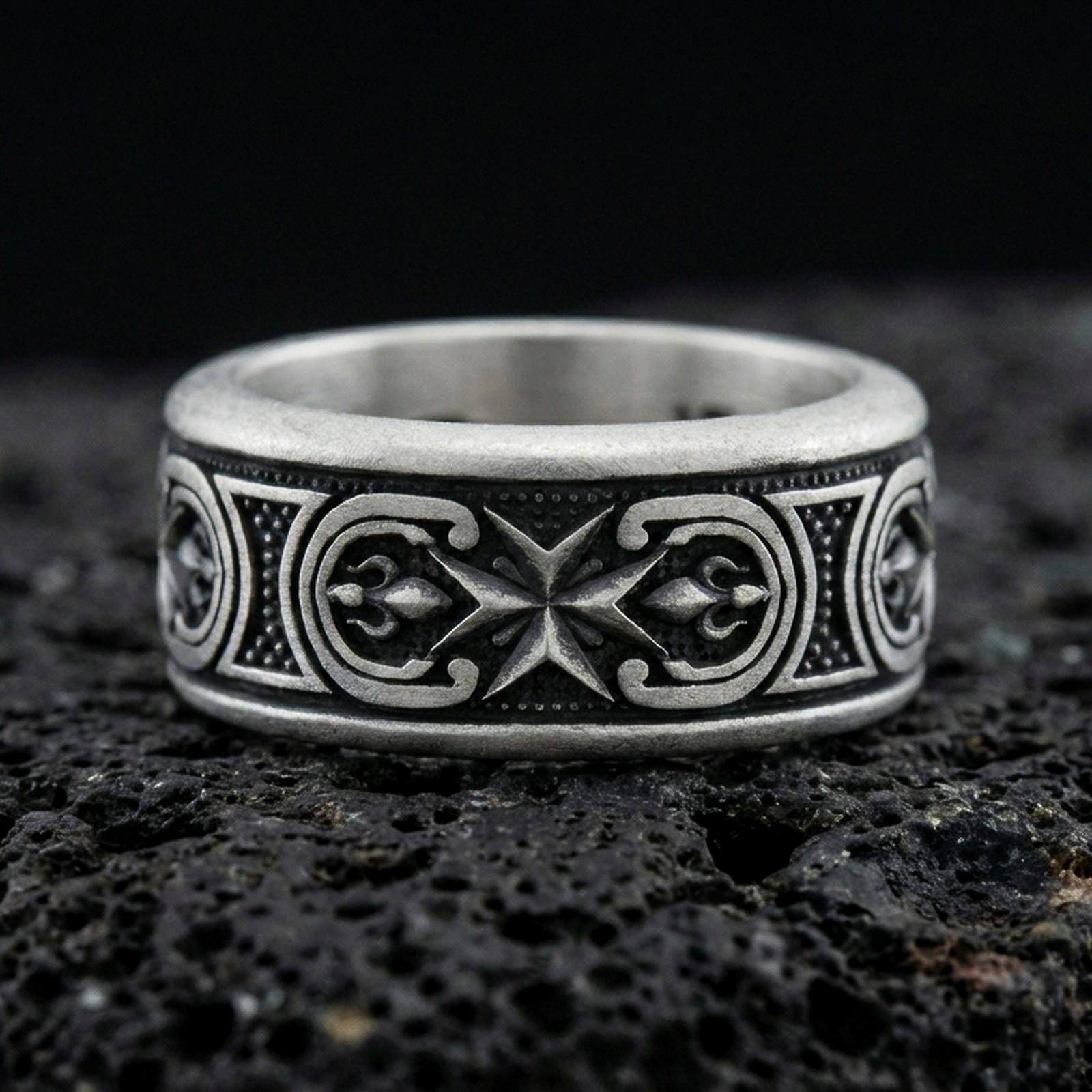 Oxidized sterling silver Maltese cross spinner band ring on dark volcanic rock, raised cross motif on rotating center with ornate side panels, premium macro product shot.