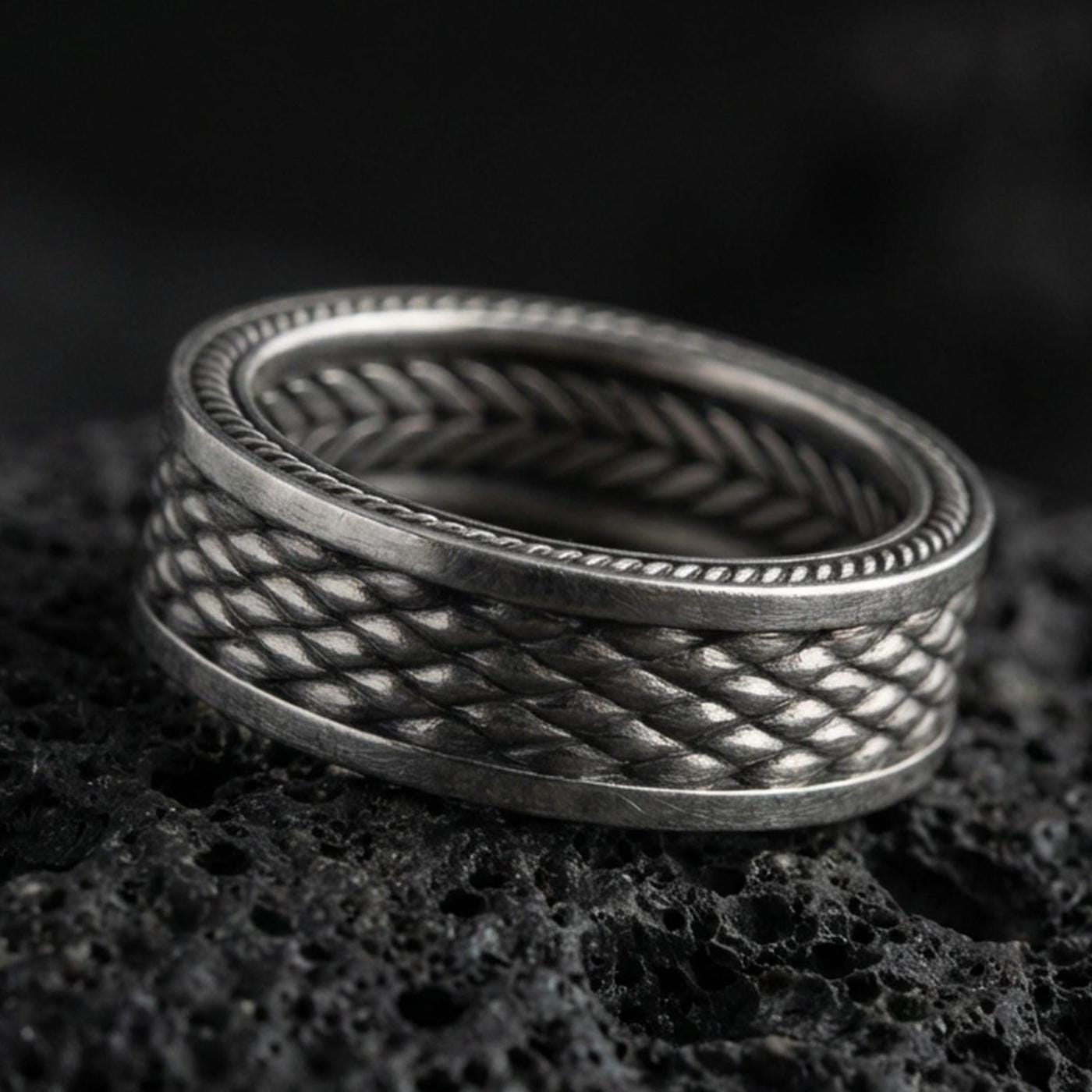 Oxidized sterling silver braided rope texture band ring on dark volcanic rock, woven pattern with dotted edge detail, premium macro product shot.