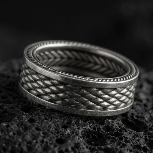 Oxidized sterling silver braided rope texture band ring on dark volcanic rock, woven pattern with dotted edge detail, premium macro product shot.
