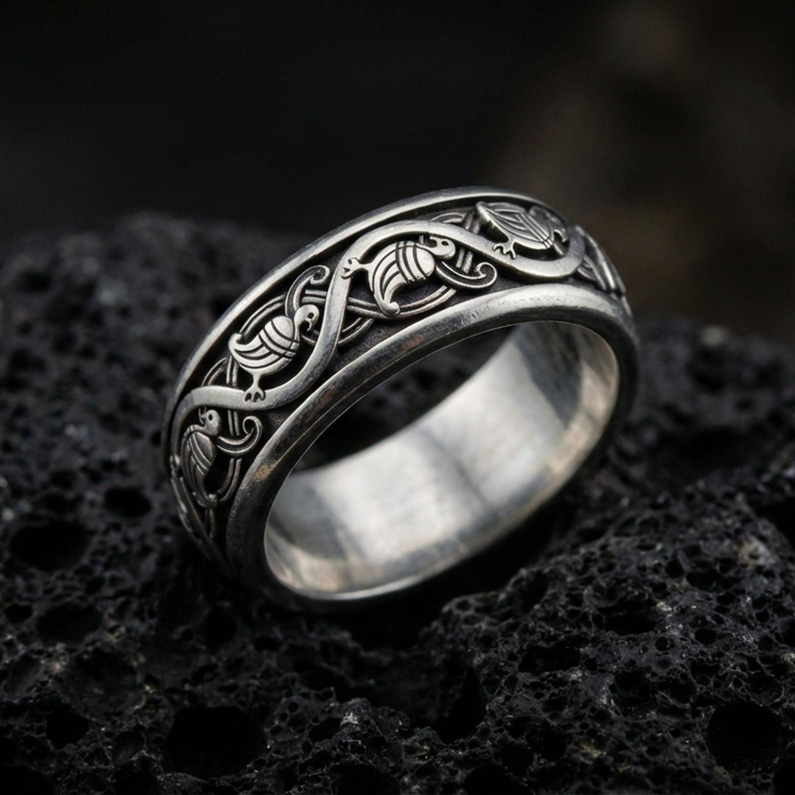 Oxidized sterling silver dove pattern band ring on dark volcanic rock, repeating bird motif with elegant scrollwork, premium macro product shot.