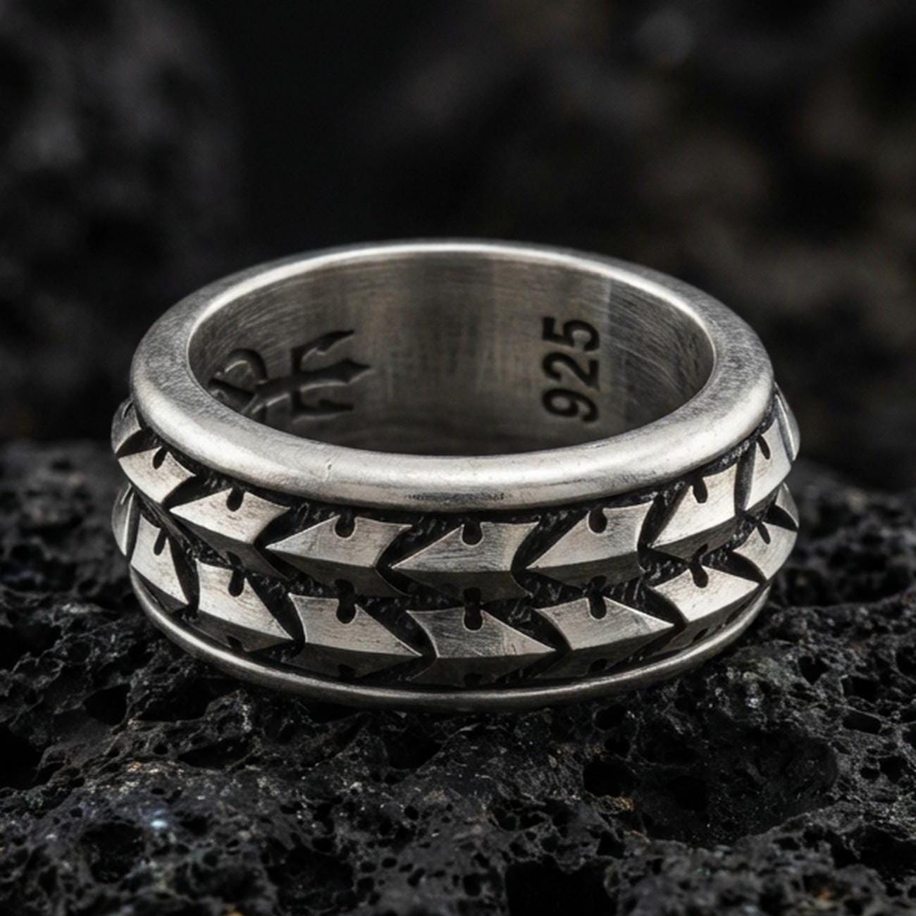 Oxidized sterling silver arrowhead spinner band ring on dark volcanic rock, tribal geometric chevron pattern on rotating center, premium macro product shot.