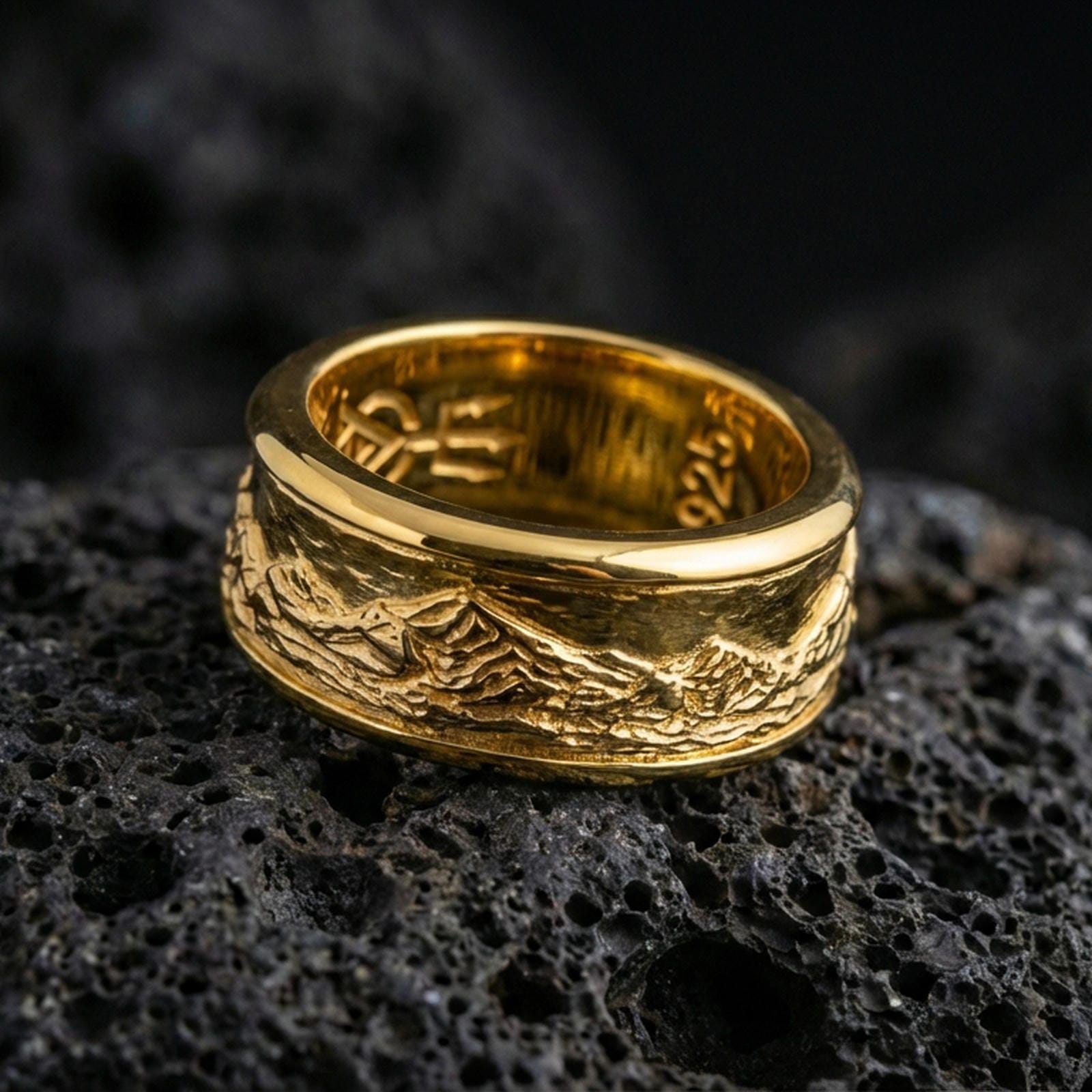 Gold plated mountain range band ring on volcanic rock, detailed alpine skyline engraving with rugged terrain texture, luxury jewelry product photography.
