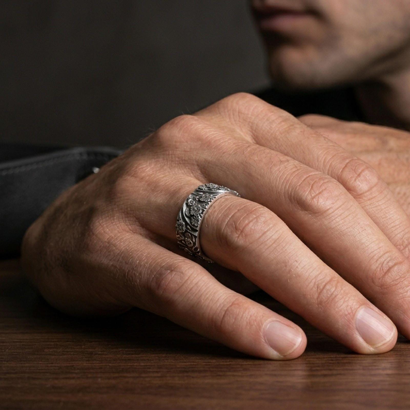 Oxidized sterling silver floral garden band worn on finger, raised daisy blossoms and leaf scroll detail, natural lifestyle hand shot with shallow depth of field.
