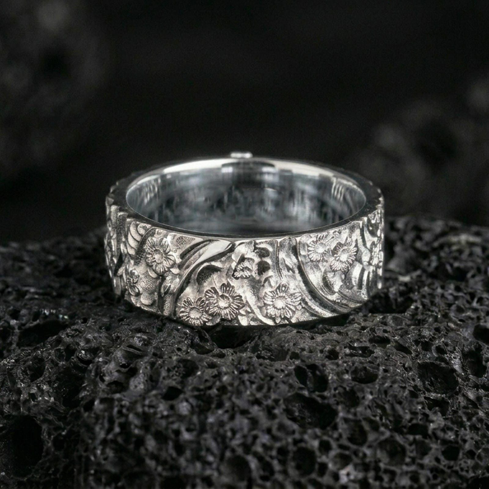 Polished silver floral engraved band ring on black volcanic stone, crisp daisy relief and swirling botanical pattern, centered close-up studio lighting.