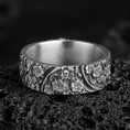 Load image into Gallery viewer, Oxidized sterling silver floral garden band ring on dark volcanic rock, raised daisy blossoms with flowing leaf scroll engraving, premium macro product shot.
