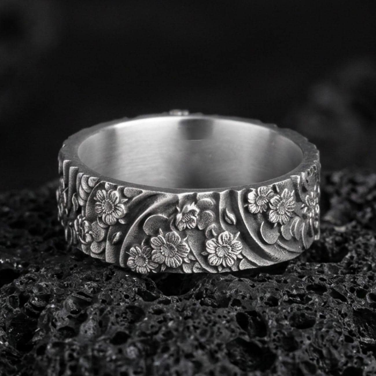 Oxidized sterling silver floral garden band ring on dark volcanic rock, raised daisy blossoms with flowing leaf scroll engraving, premium macro product shot.