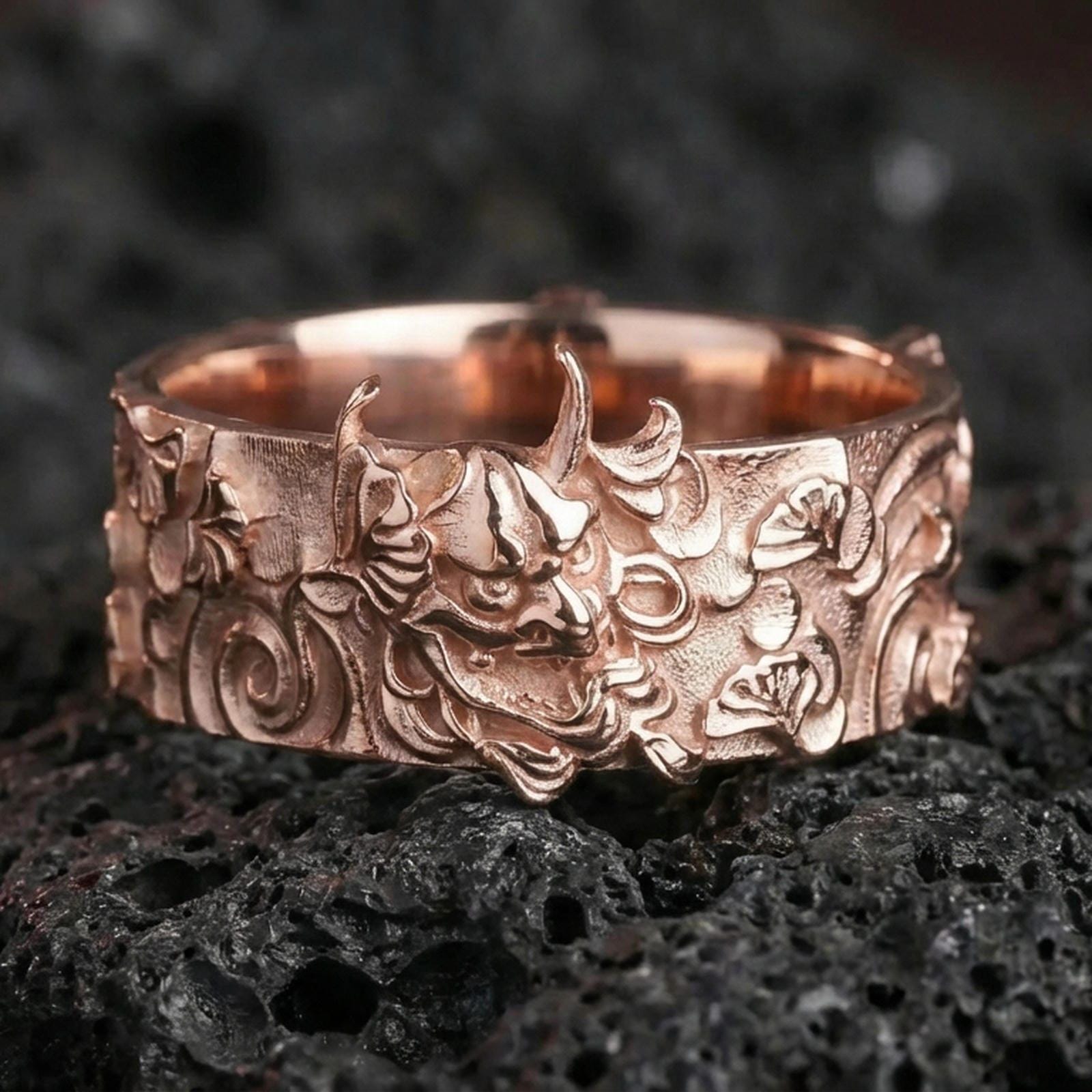 Rose gold plated sterling silver oni mask ring on volcanic stone, sculpted demon face relief with deep contrast and ornate side motifs, minimal high-end close-up.