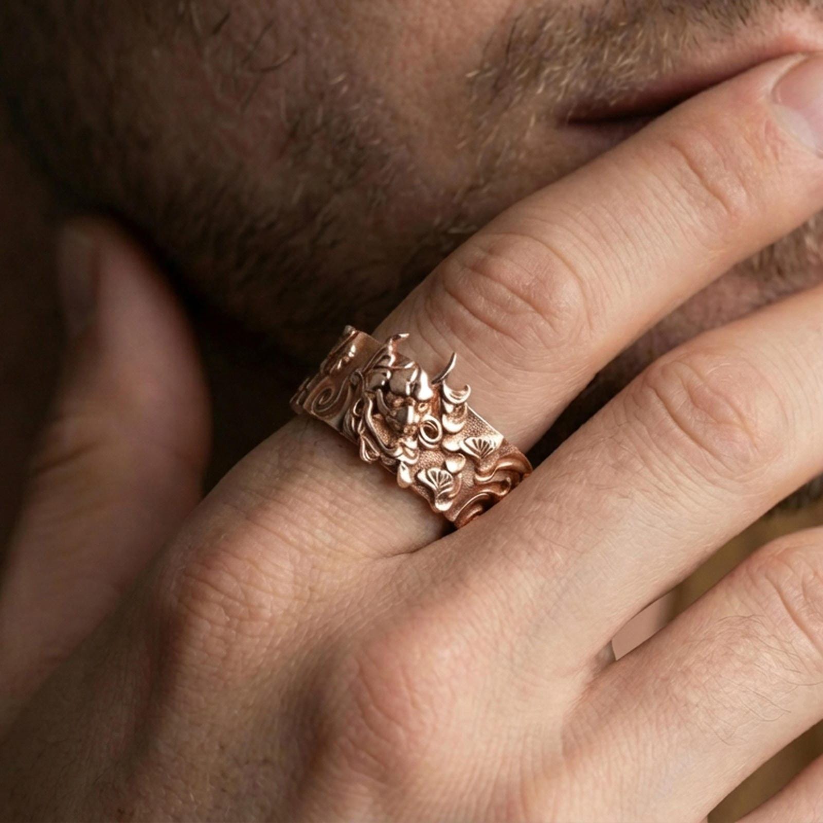 Rose gold plated oni mask ring worn on finger, sculpted demon face relief with ornate side motifs, elegant lifestyle wear shot.