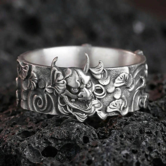 Oxidized sterling silver oni mask band ring on dark volcanic rock, Japanese demon face relief with swirling cloud motifs, premium macro product shot.