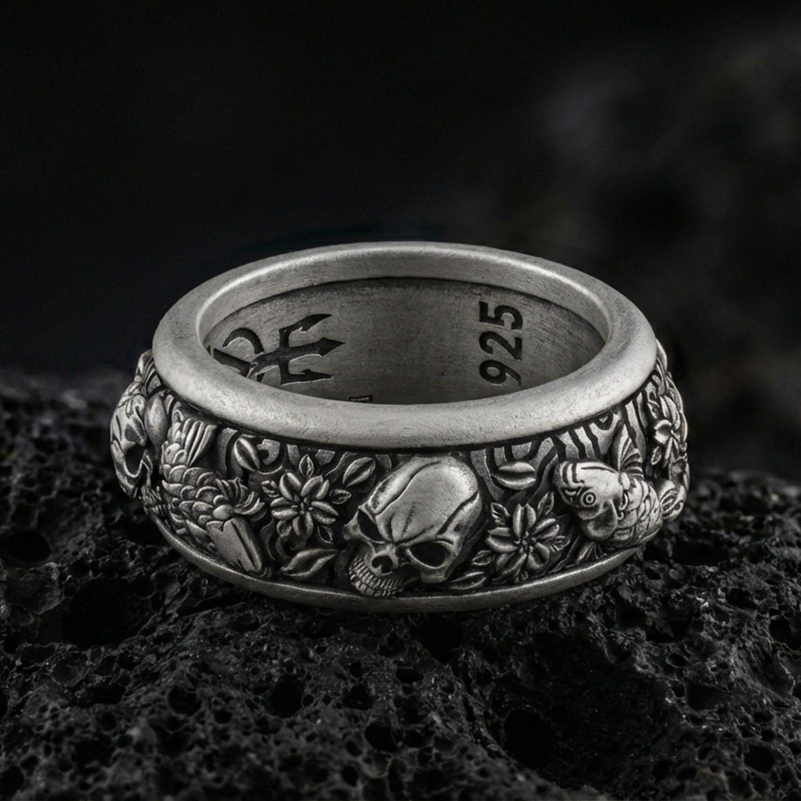 Oxidized sterling silver skull and koi spinner band ring on dark volcanic rock, detailed skull relief with carp, clouds, and wave motifs on rotating center, premium macro product shot.