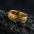 Load image into Gallery viewer, Gold plated tree roots band ring on volcanic rock, detailed branch and leaf engraving with organic texture, luxury jewelry product photography.

