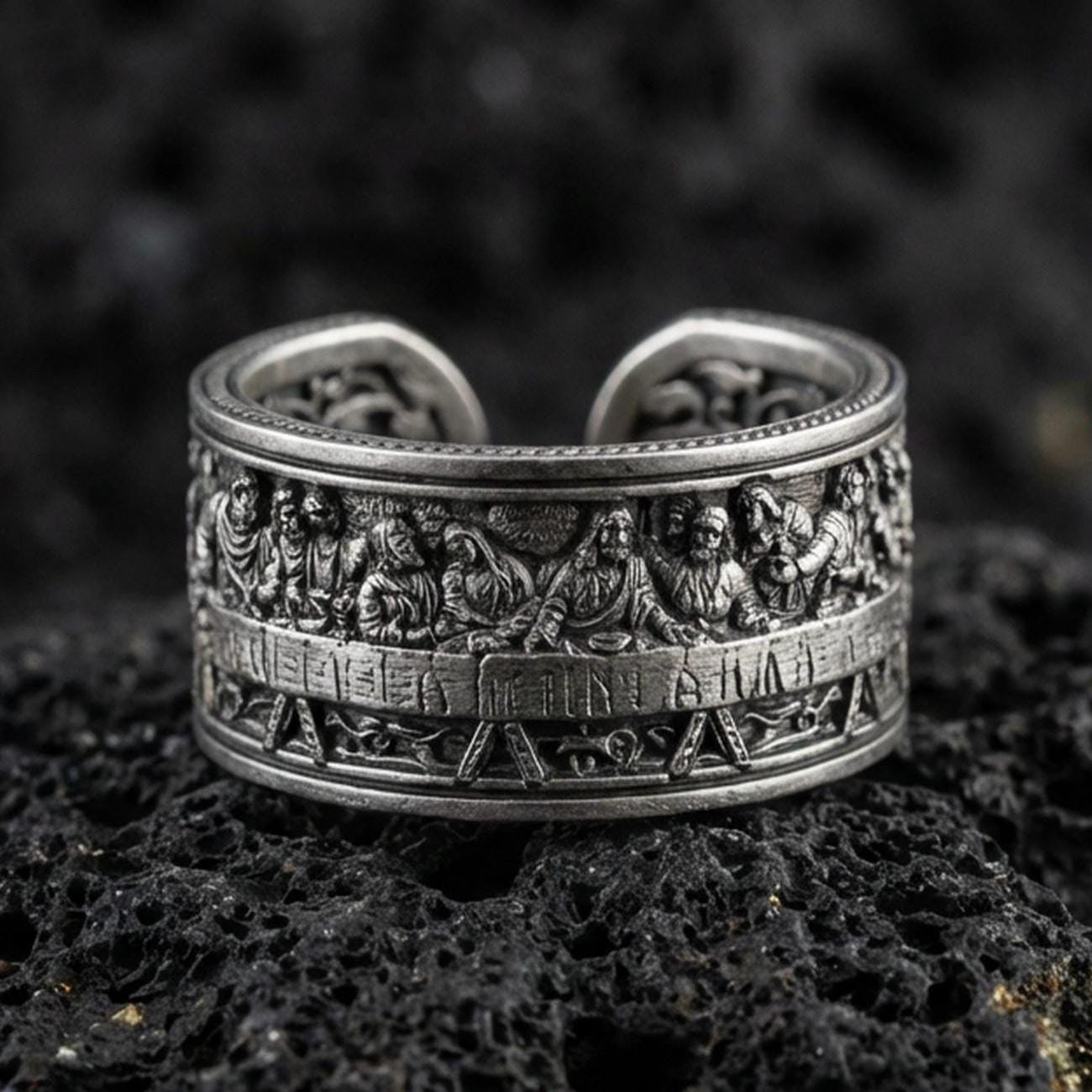 Oxidized sterling silver Last Supper engraved band ring on dark volcanic rock, high-relief apostle scene with detailed table setting, premium macro product shot.