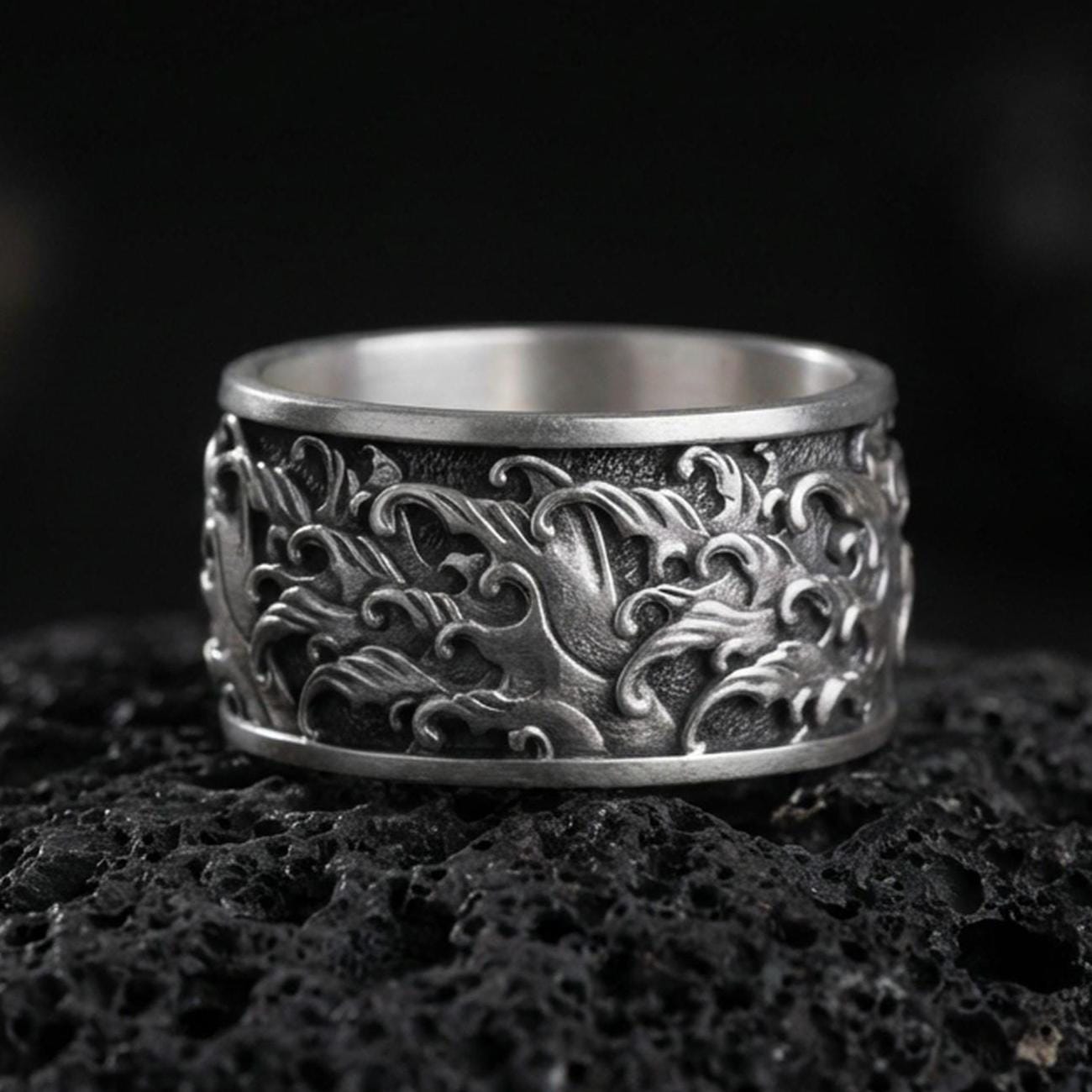 Oxidized sterling silver swirling flames band ring on dark volcanic rock, high-relief fire wave engraving with deep contrast, premium macro product shot.